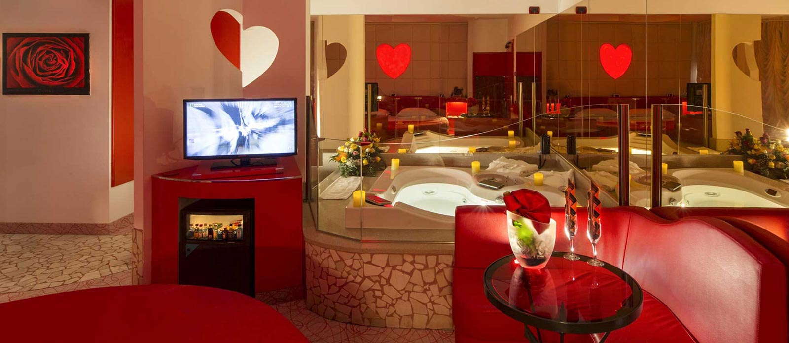 Motel K Junior Suite Cupido vista dal letto rotondo, TV orientabile verso il letto, vasca idromassaggio a forma di cuore, bottiglia di champagne in secchiello del ghiaccio appoggiato sul tavolo assieme a 2 bicchiere con servizio candele in suite camera a tema Amore