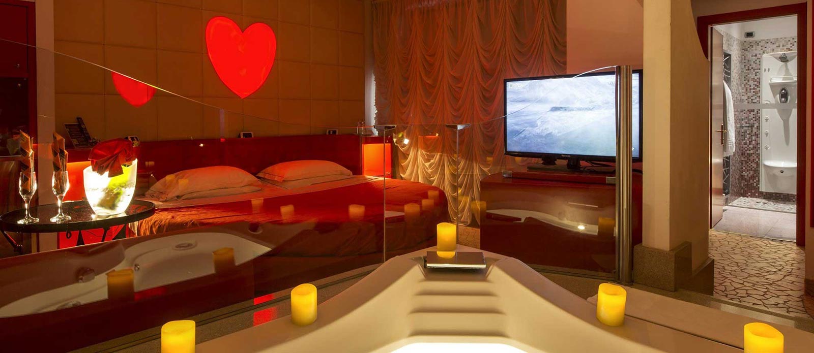 Motel K Junior Suite Cupido vista dalla vasca idromassaggio TV orientabile verso la vasca, letto rotondo, bottiglia di champagne in secchiello del ghiaccio appoggiato sula tavolo assieme a 2 bicchiere con servizio candele in suite camera a tema Amore