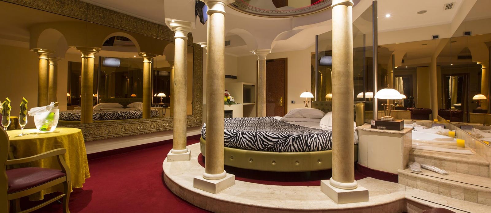 Motel K Senior Suite Impero Motel K Senior Suite Impero vista sul letto rotondo matrimoniale circondato da colonne di marmo e giochi di specchio, vasca idromassaggio doppia dietro al letto, bottiglia di champagne in secchiello del ghiaccio appoggiata sul tavolo assieme a 2 bicchieri, camera a tema Impero Romano